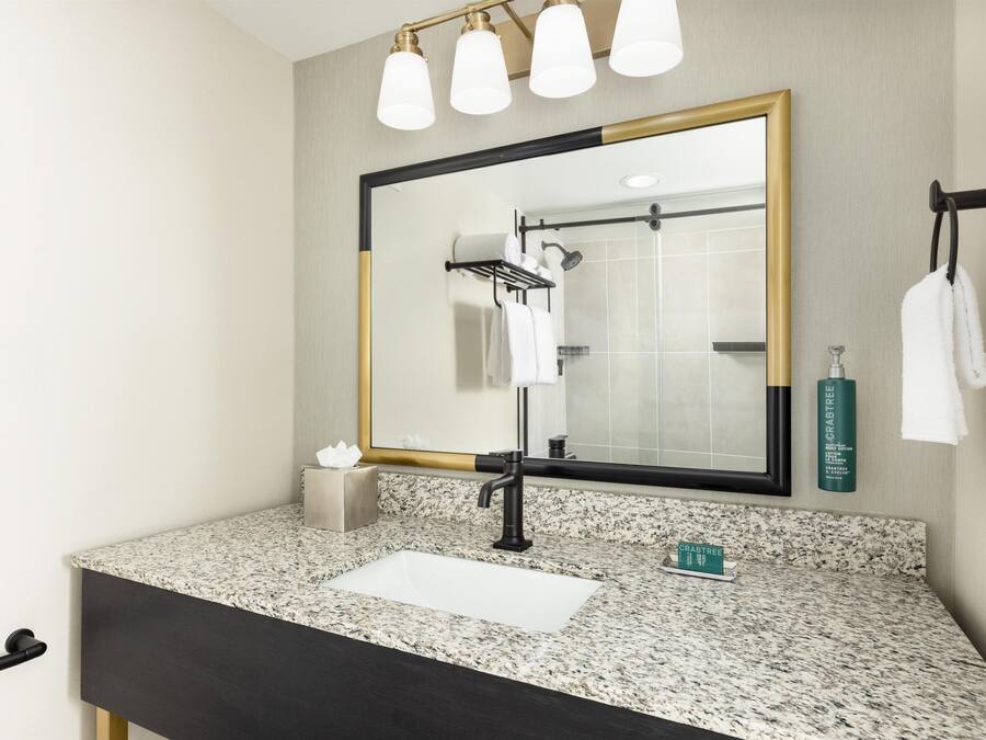 A bathroom with a granite countertop, a black sink, a mirror with a gold frame, a towel rack, and a shower.