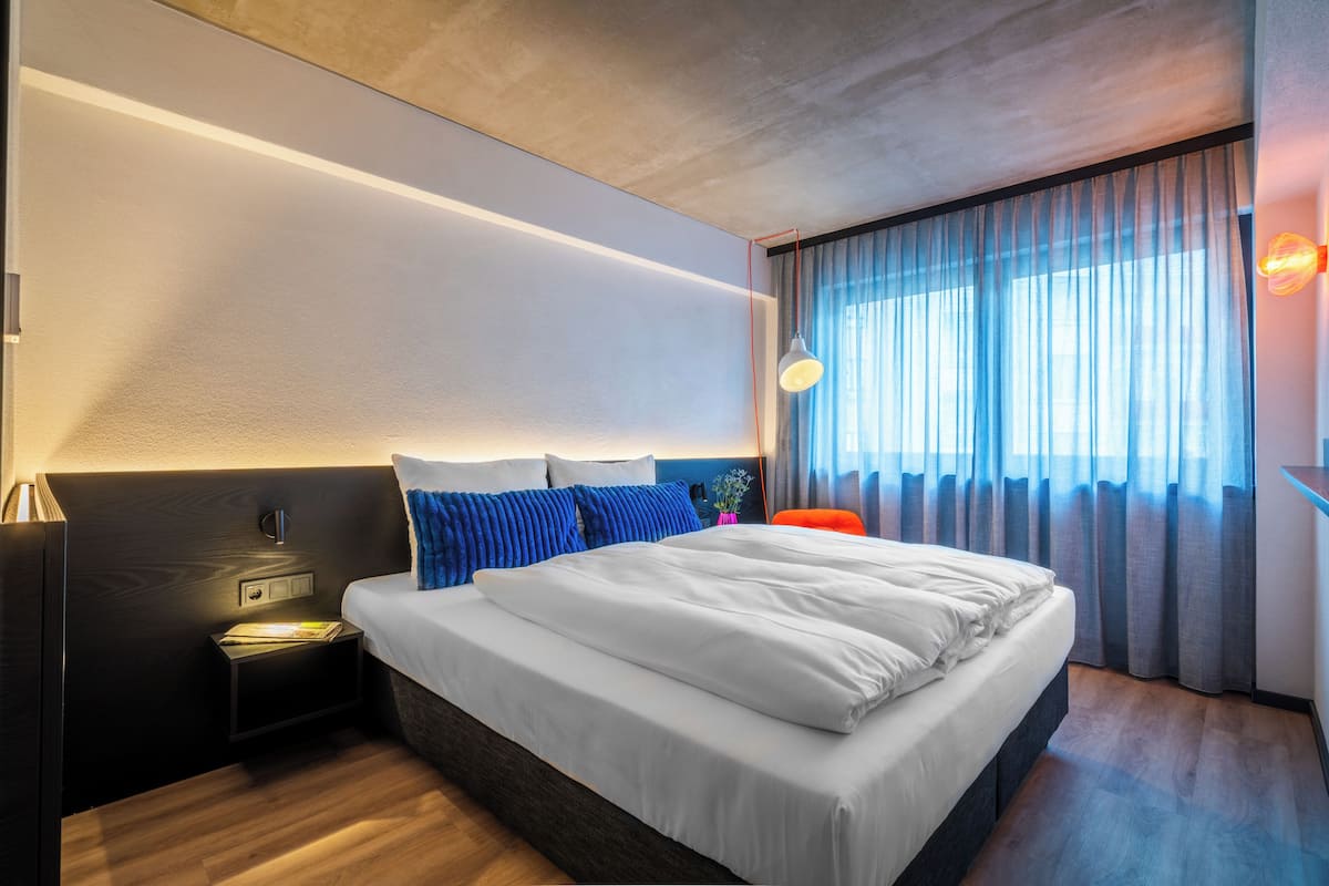 Une chambre d’hôtel moderne avec un grand lit, des tables de chevet, une petite table, une lampe et une fenêtre avec des rideaux.