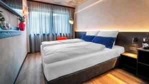 Ein modernes Hotelzimmer mit einem großen Bett, einem Nachttisch, einem Beistelltisch, einem roten Sessel und einem Fenster mit Vorhängen.