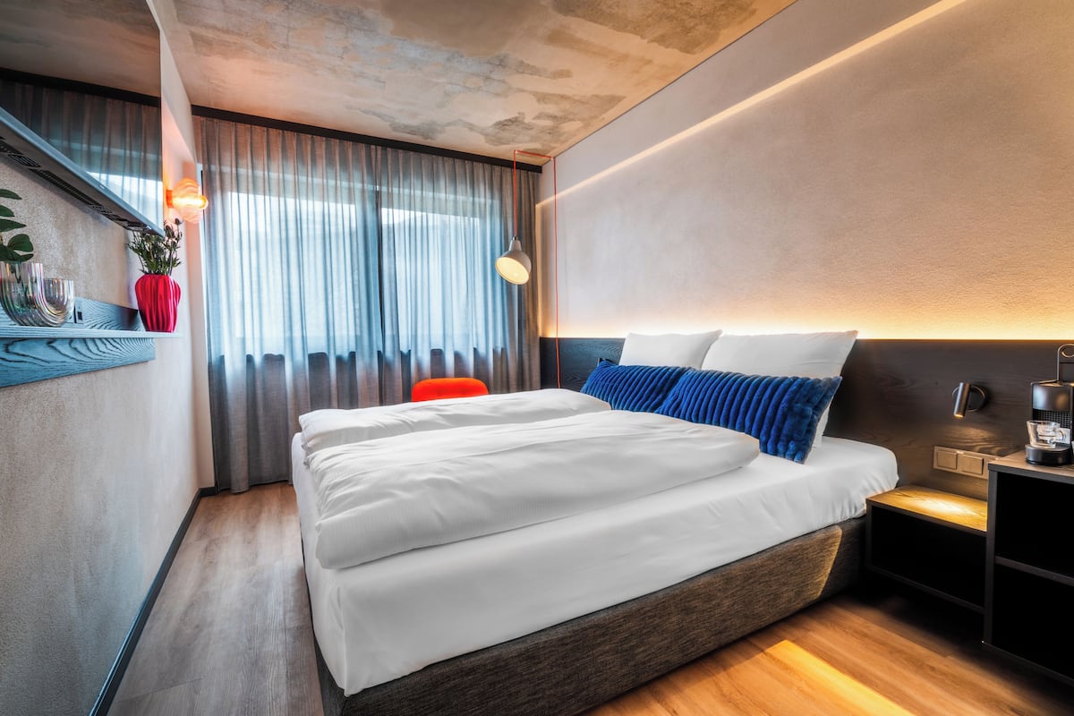 Une chambre d’hôtel moderne avec un grand lit, une table de chevet, une commode, un fauteuil rouge et une fenêtre avec des rideaux.