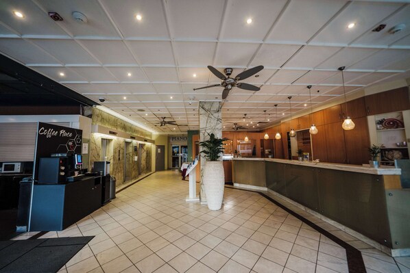 A hotel lobby with a reception desk, a coffee machine, a piano, and a large vase with a plant.