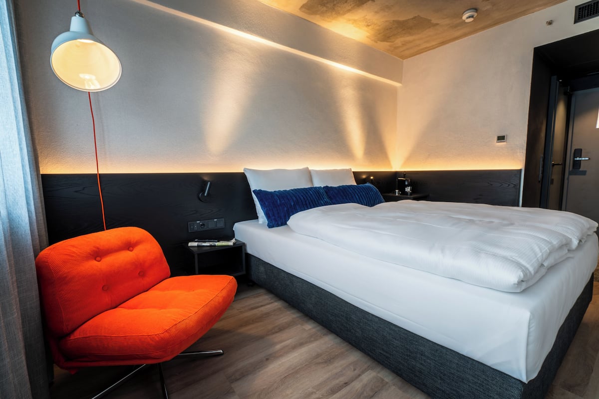 Une chambre d’hôtel moderne avec un grand lit, une table de chevet, un fauteuil rouge et une lampe suspendue.