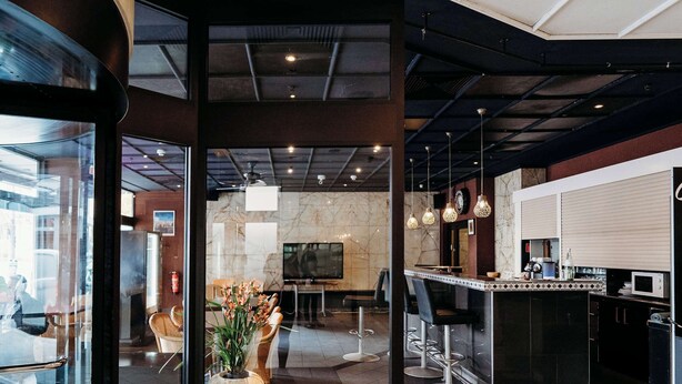 Eine moderne Hotelhalle mit Glastür, einer zentralen Sitzecke mit Pflanze, einer Bar mit Hockern und einer Küchenzeile.