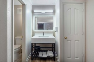 Un baño estrecho con un lavabo blanco, un espejo encima y un estante negro debajo.