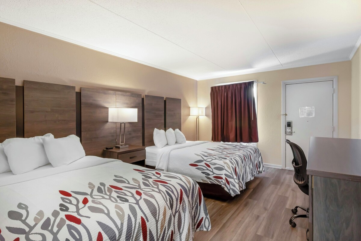 A hotel room with two beds, a desk, a chair, a lamp, and a door.