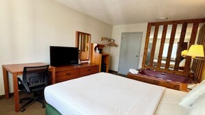 A hotel room with a bed, a desk, a chair, a television, a mirror, a bathroom with a sink, and a door. - Americas Best Value Inn St. Ignace (St Ignace)