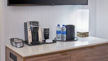 Habitación de hotel con kitchenette moderna, cafetera, dos botellas de agua, un cubo de basura y un televisor de pantalla plana montado en la pared.