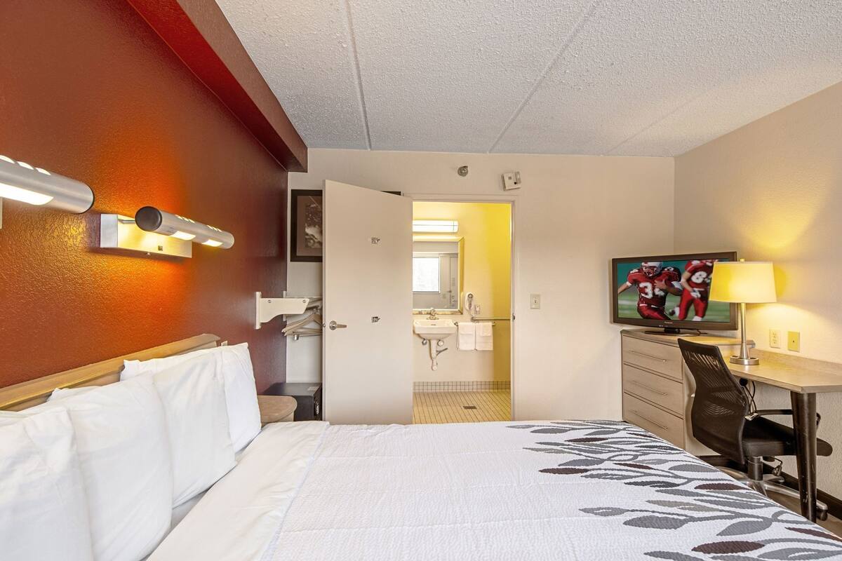 A hotel room with a bed, a desk with a chair, a television, and a bathroom visible through an open door.