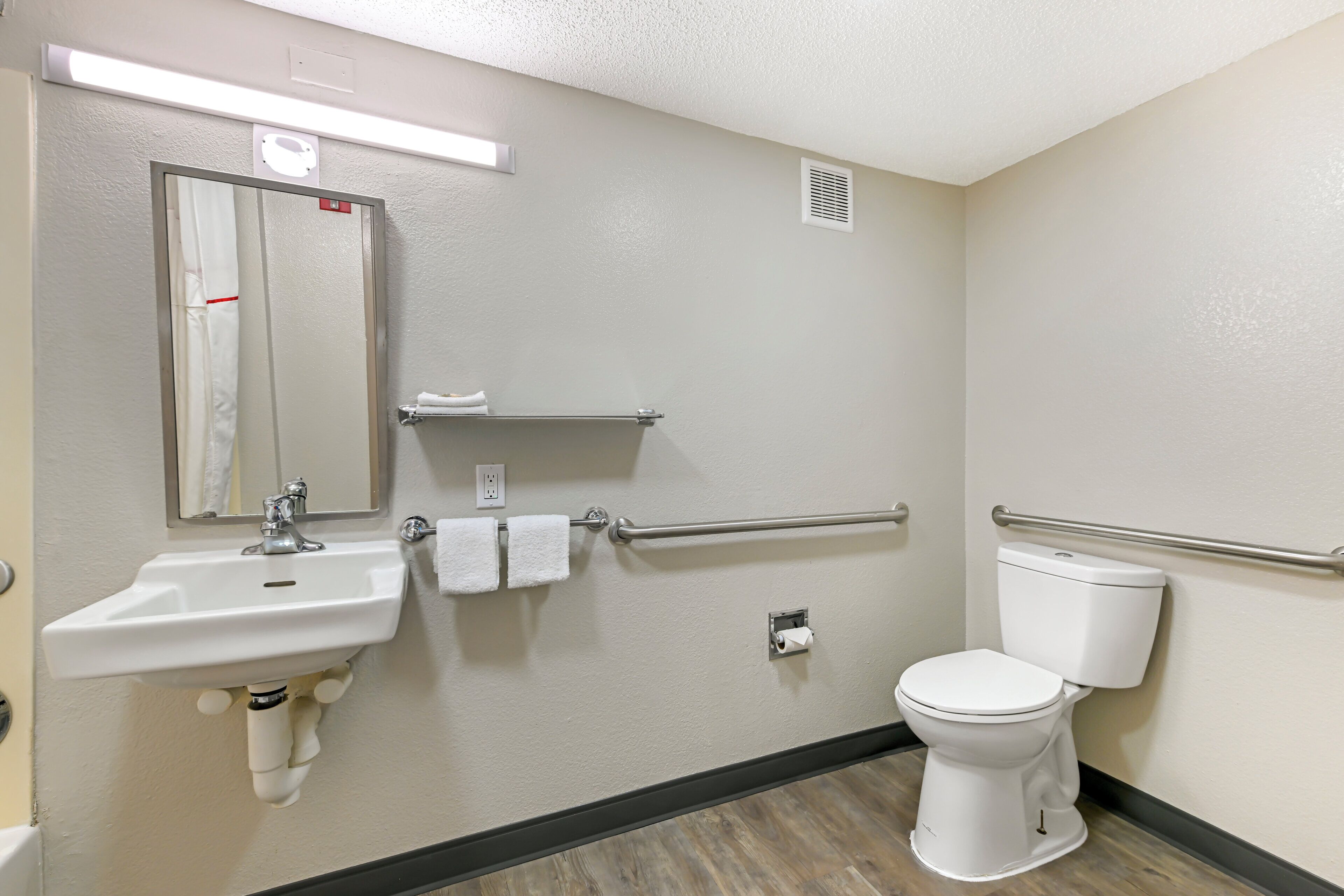 A bathroom with a sink, mirror, towel rack, toilet, and handrail.
