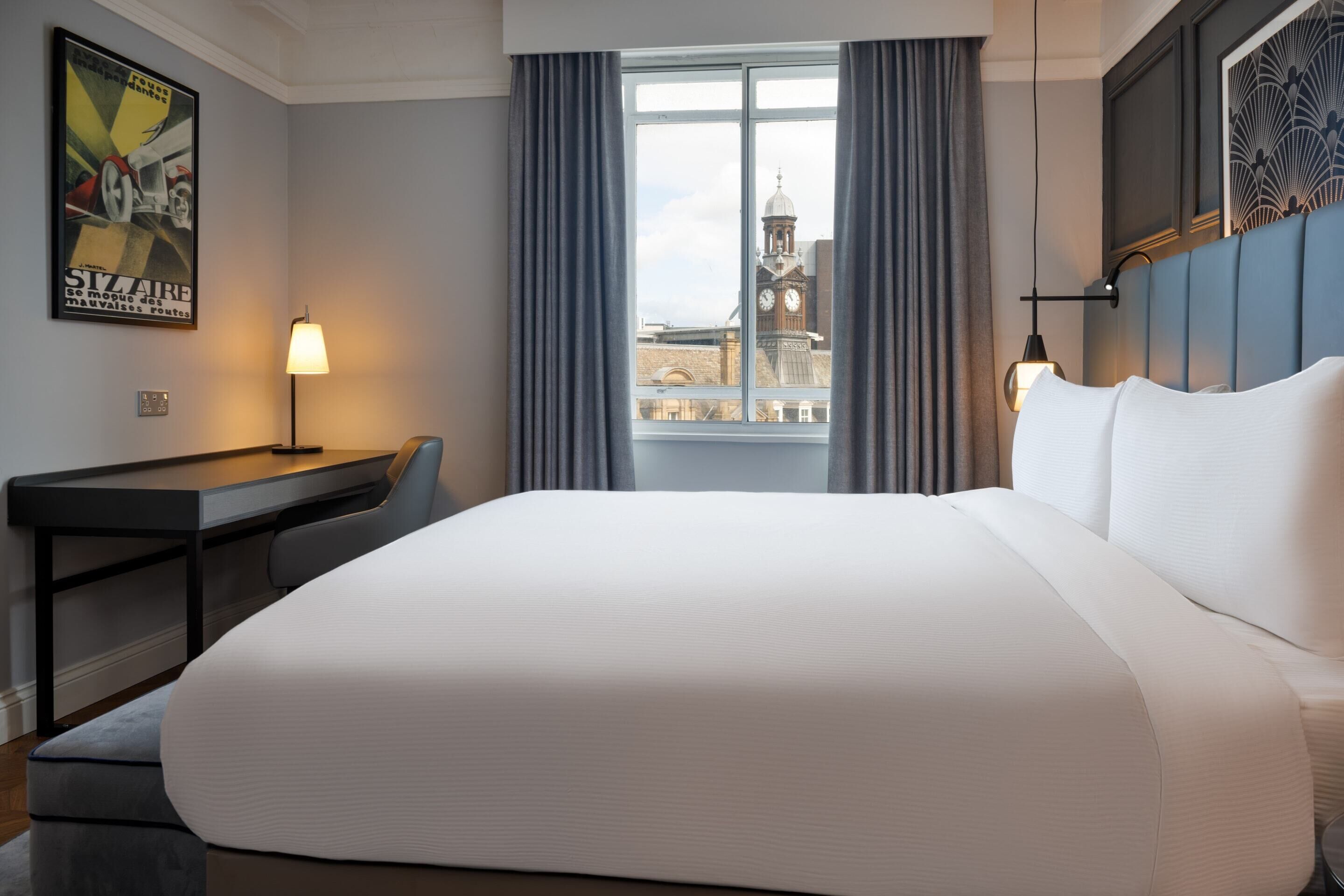 Een hotelkamer met een groot bed, een bureau, een stoel, een lamp en een raam met uitzicht op een klokkentoren.