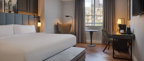 A hotel room with a large bed, a desk with a chair, a small table, and a window with curtains.