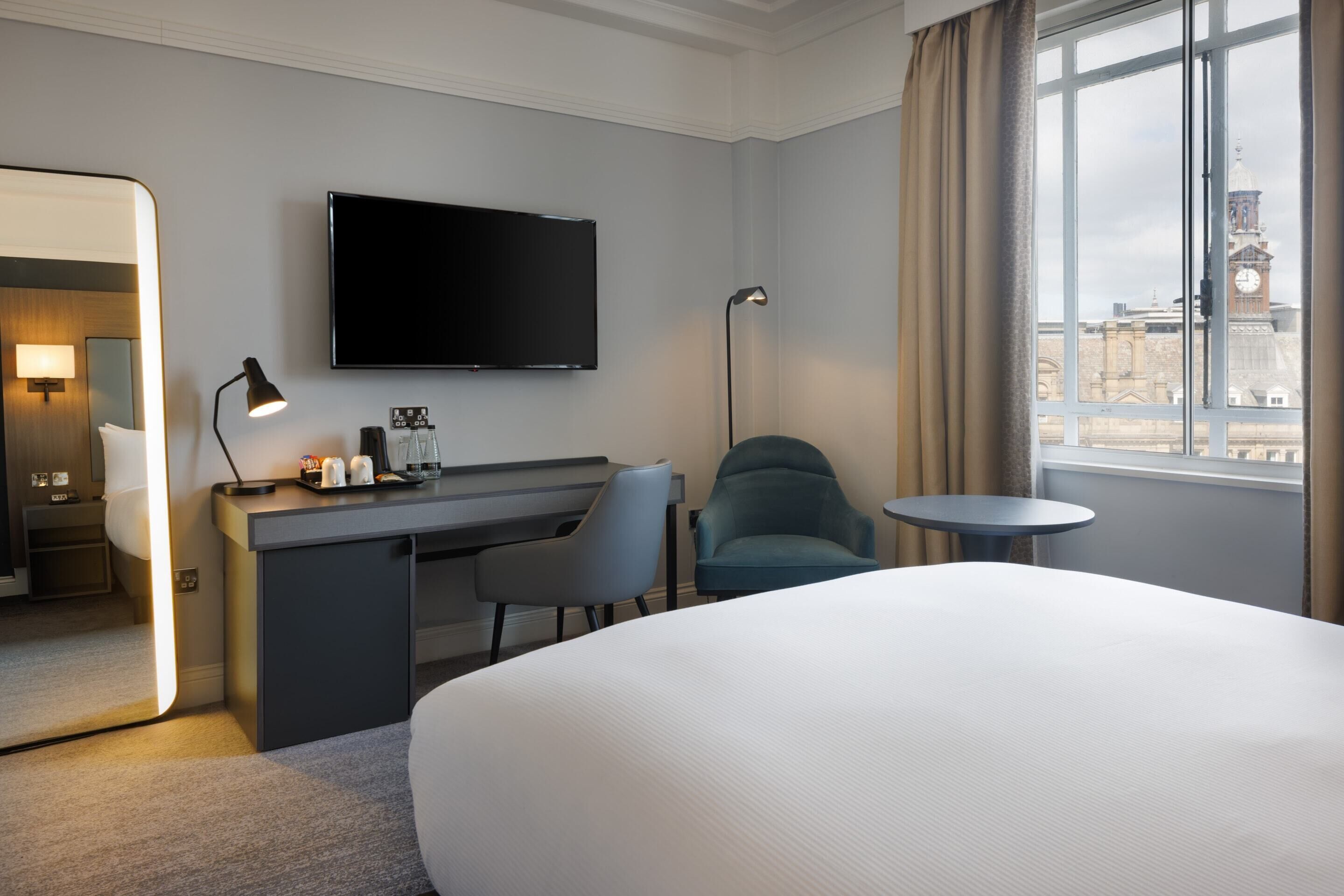 A hotel room with a bed, desk, chair, TV, lamp, and a view of a clock tower.