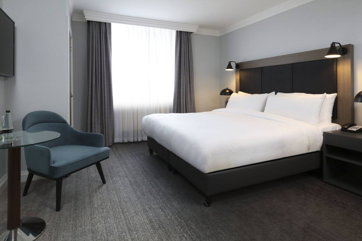 Une chambre d’hôtel avec un grand lit, une chaise, une table de chevet avec une lampe et un téléviseur fixé au mur.