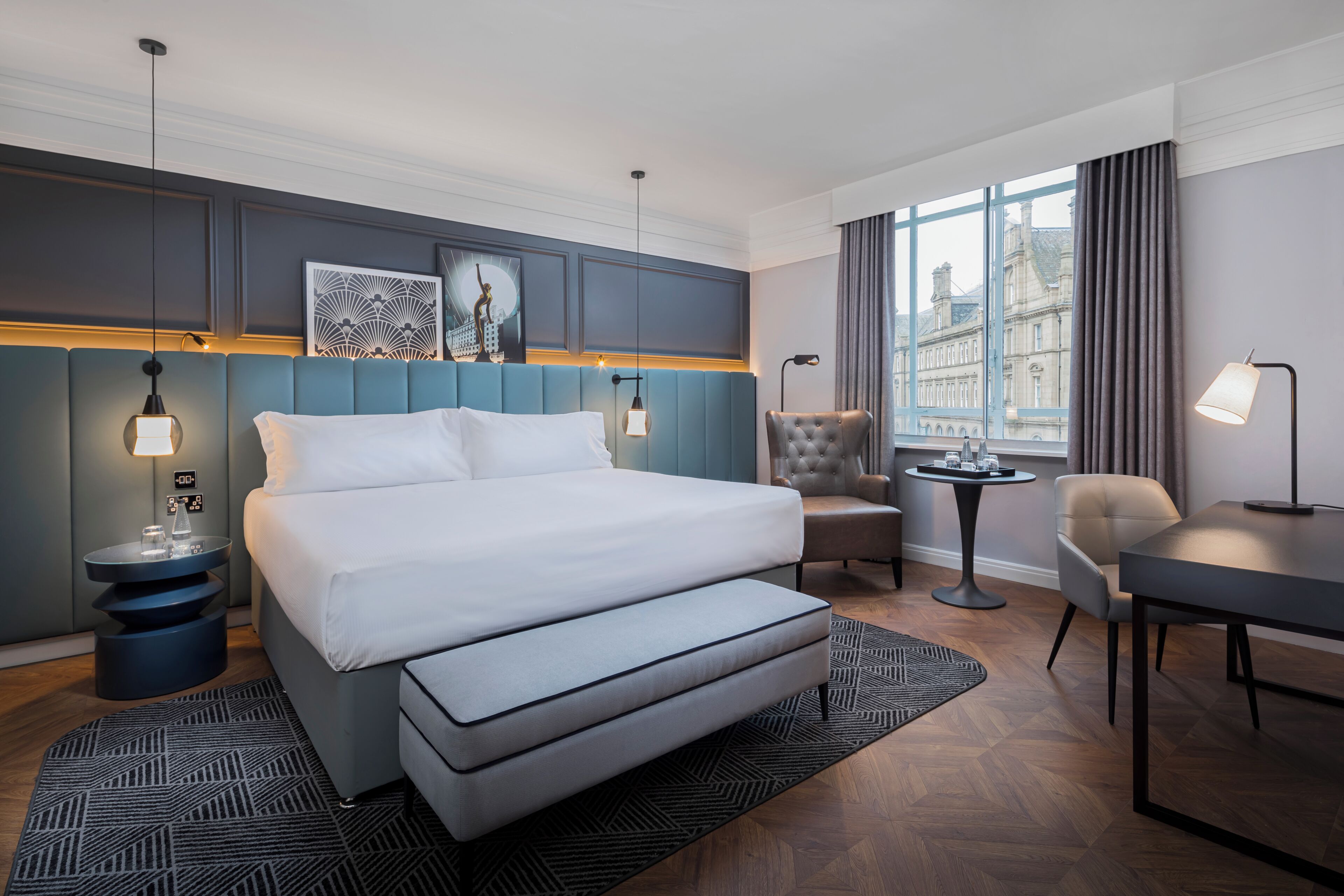A hotel room with a bed, a chair, a table, and a view of a building through the window.