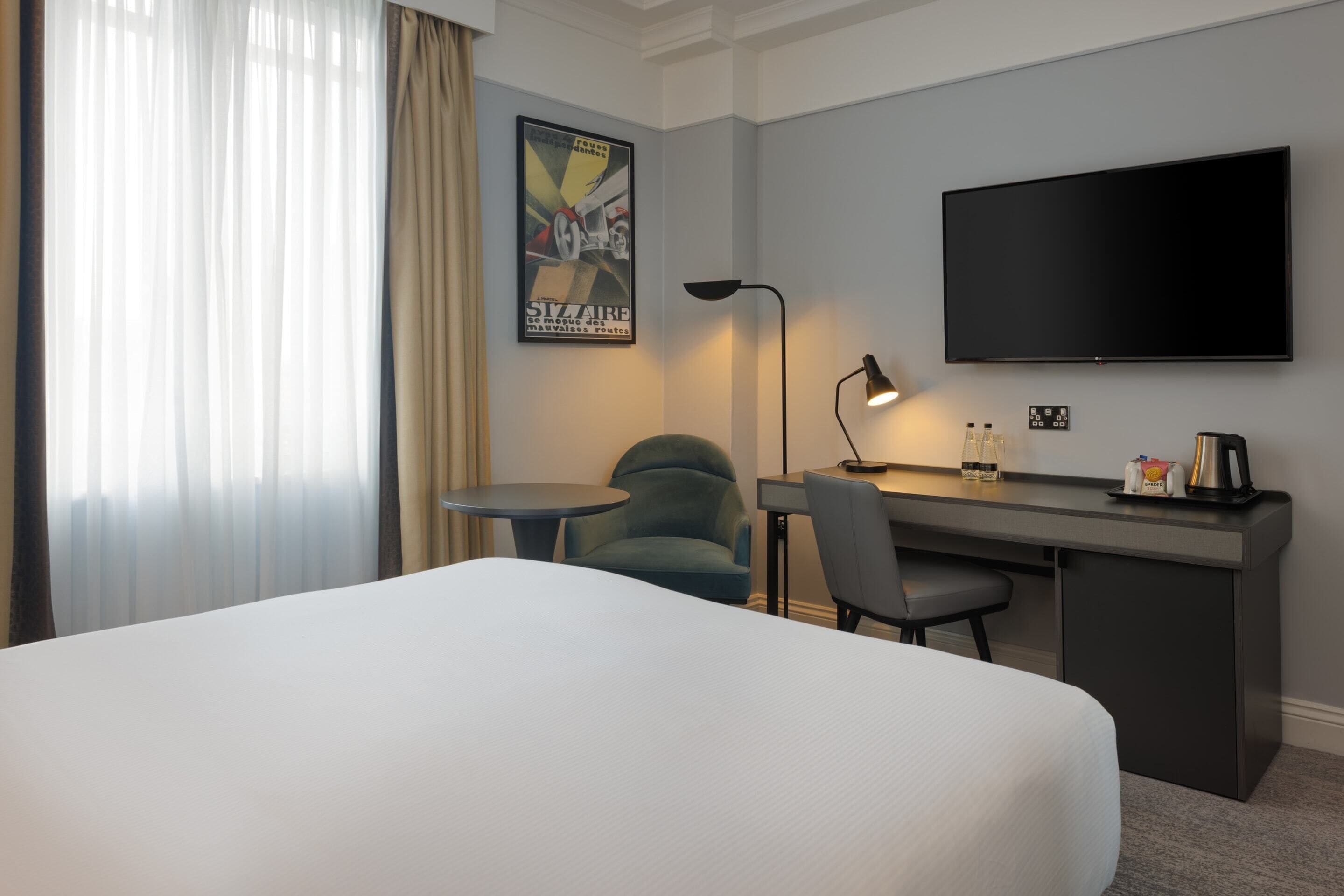 Una habitación de hotel con una cama, un escritorio, una silla, un televisor, una lámpara y un cartel en la pared.