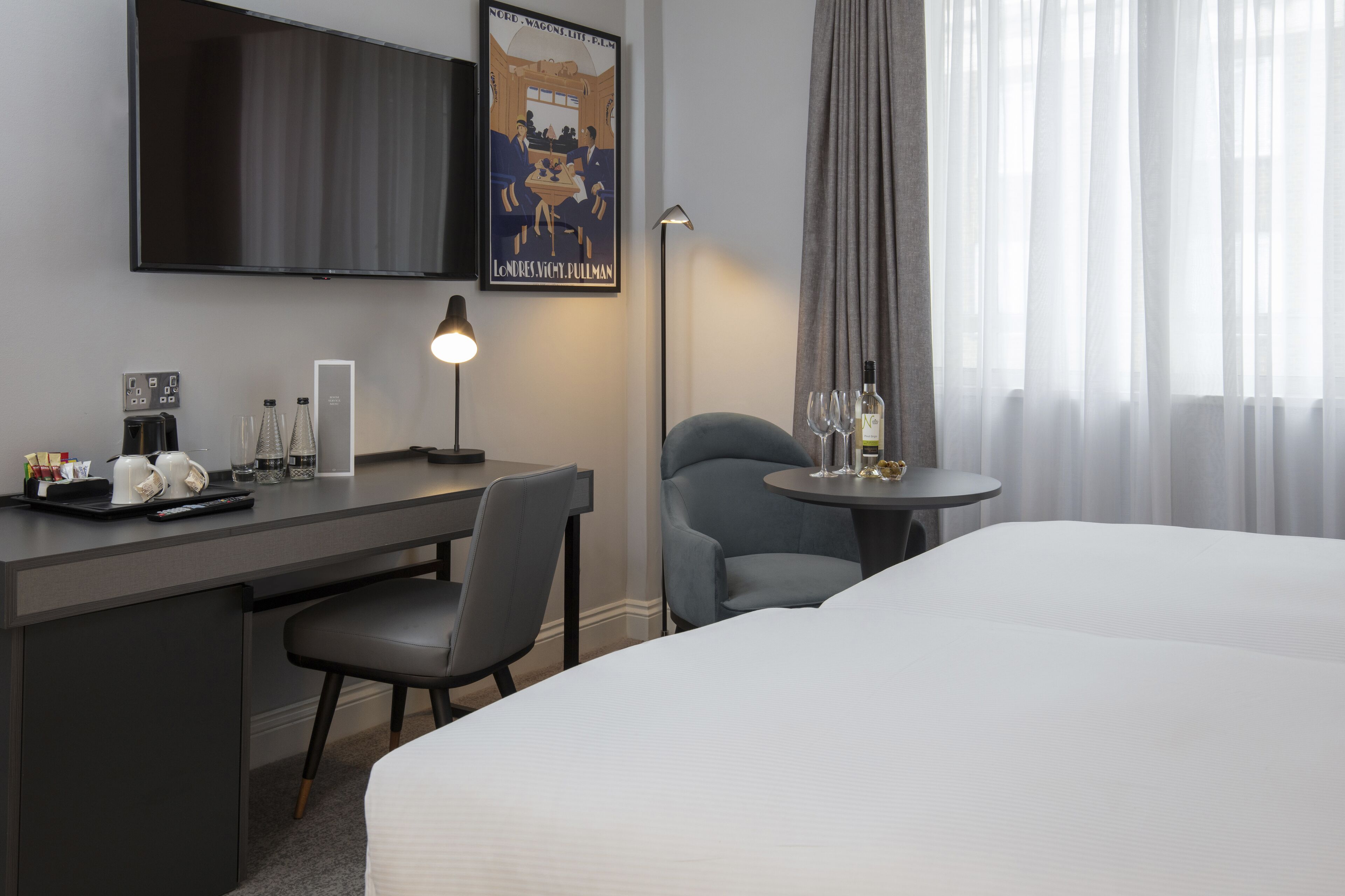 A hotel room with a bed, a desk with a chair, a small table with wine glasses, a lamp, and a poster on the wall.