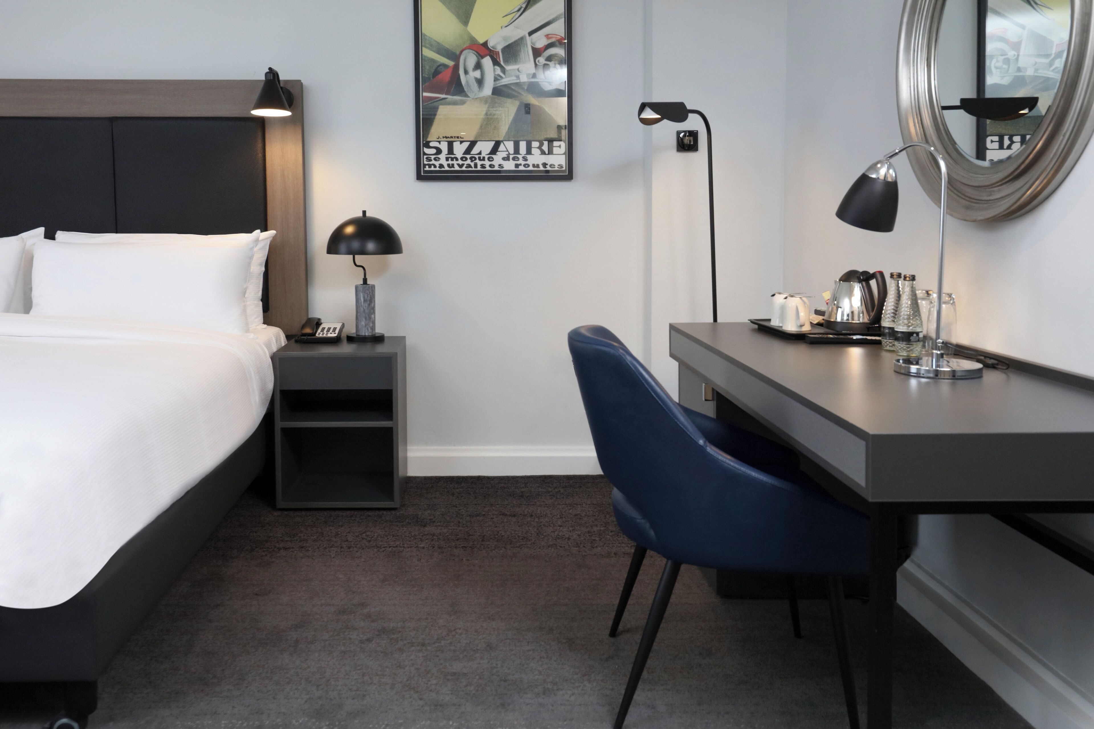 Habitación de hotel con cama, escritorio, silla, mesita de noche, lámpara, espejo y un cartel en la pared.