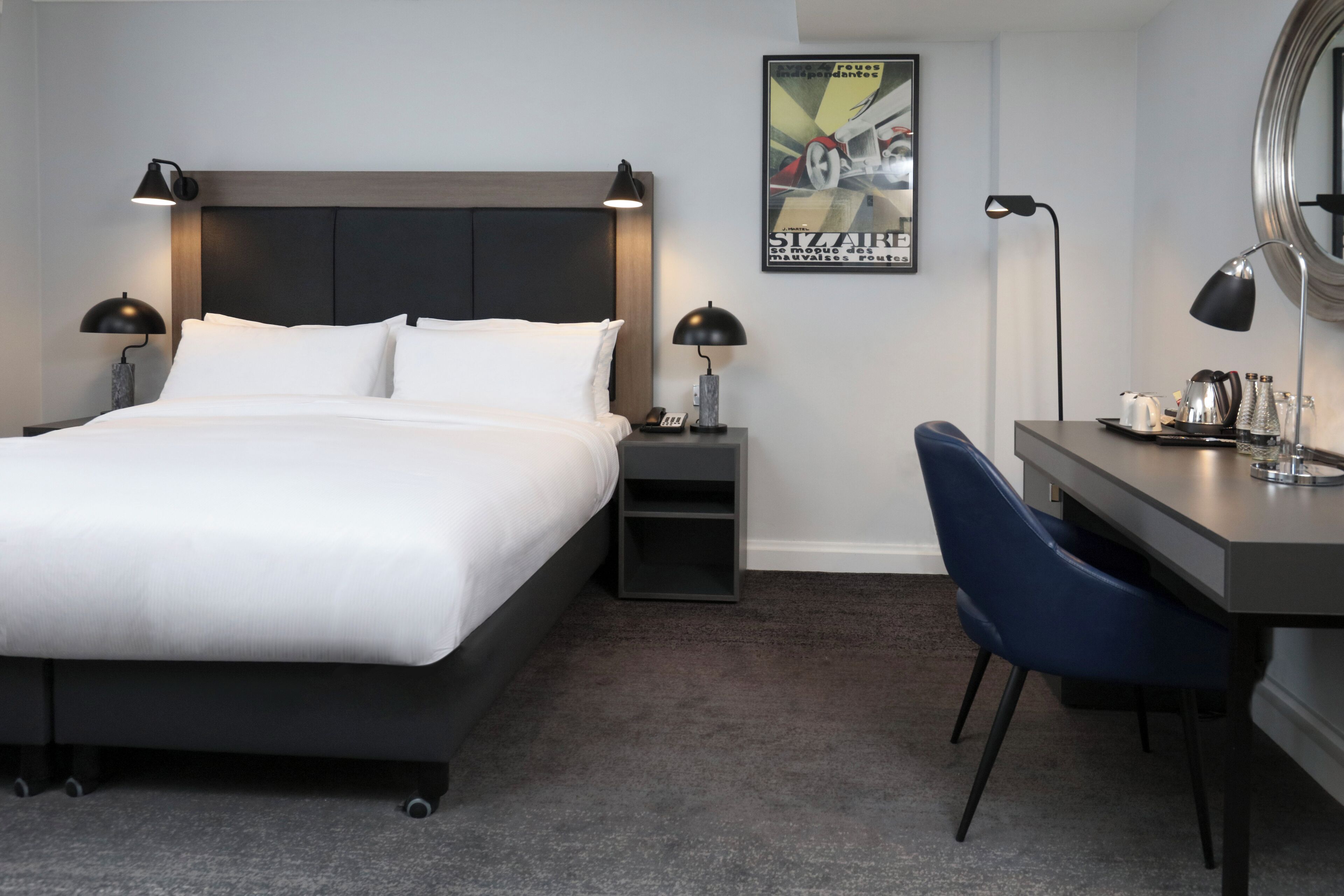 A modern hotel room with a large bed, bedside tables with lamps, a desk with a chair, and a poster on the wall.