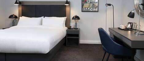 A modern hotel room with a large bed, bedside tables with lamps, a desk with a chair, and a poster on the wall.
