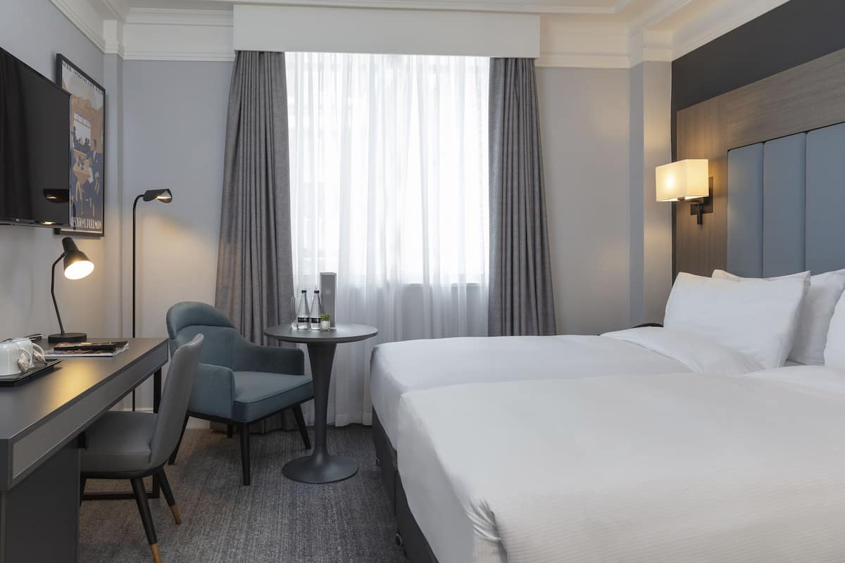Une chambre d’hôtel avec un grand lit, un bureau avec une chaise, une petite table, une télévision et une fenêtre avec des rideaux.