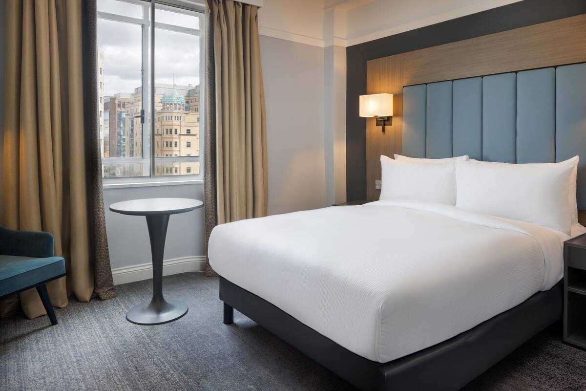 Une chambre d’hôtel avec un grand lit, une petite table ronde, une chaise bleue, une fenêtre avec des rideaux et une vue sur la ville.