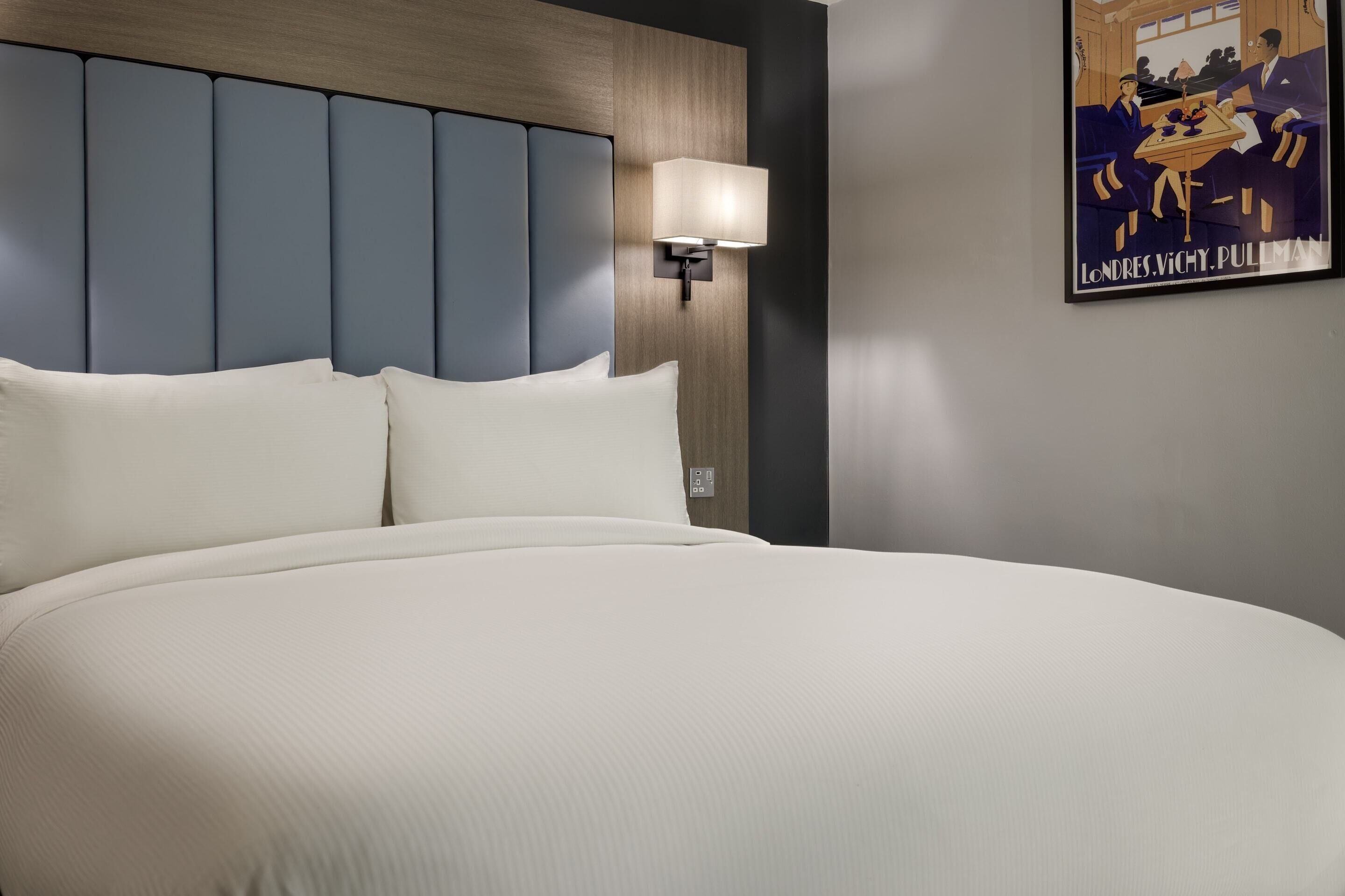 A large bed with a headboard, a wall-mounted lamp, and a poster on the wall.