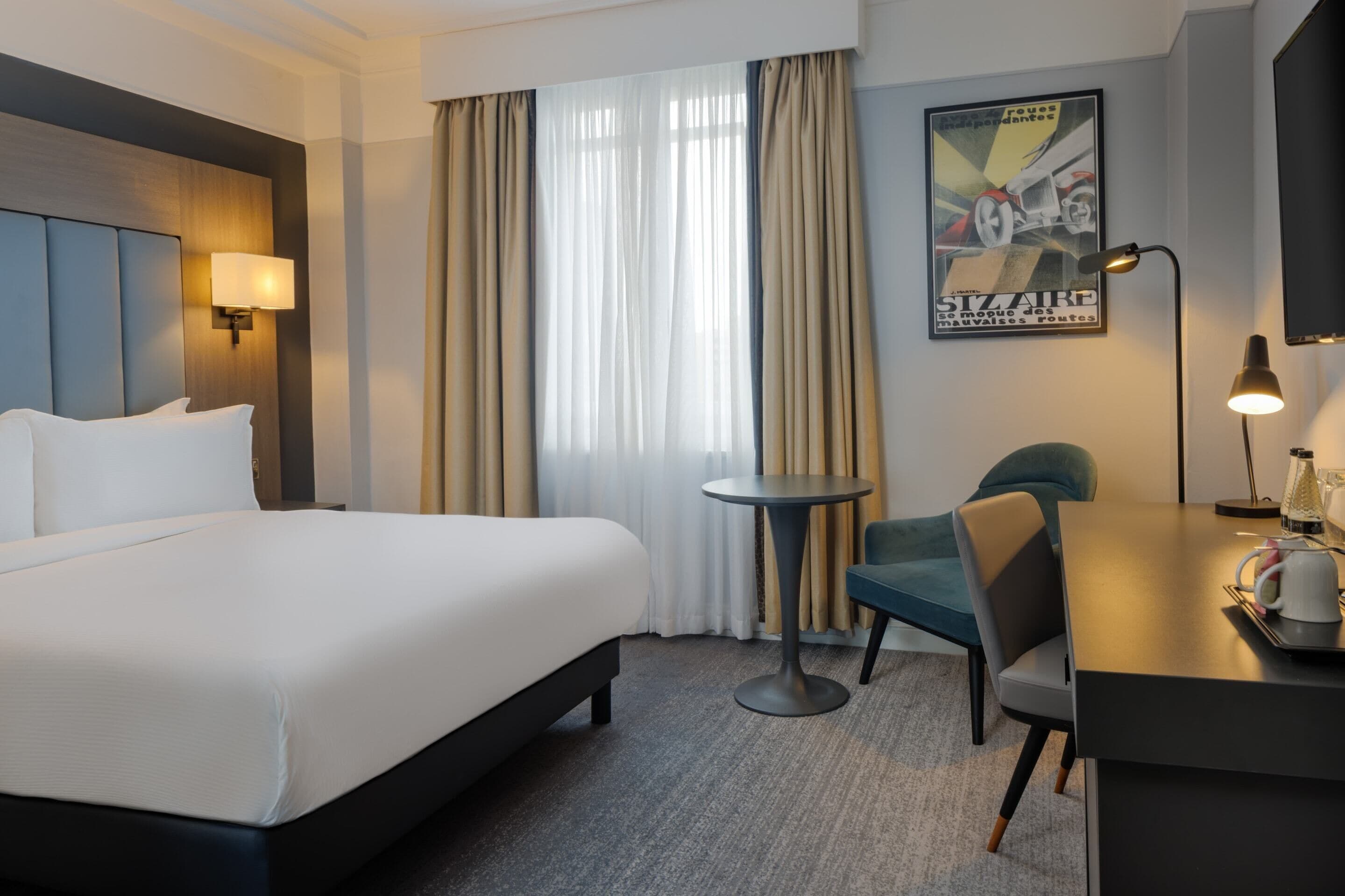 Habitación de hotel con una cama grande, una mesita redonda pequeña, una silla, un escritorio con una lámpara y un cartel en la pared.