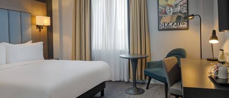 Une chambre d’hôtel avec un grand lit, une petite table ronde, une chaise, un bureau avec une lampe et une affiche accrochée au mur.