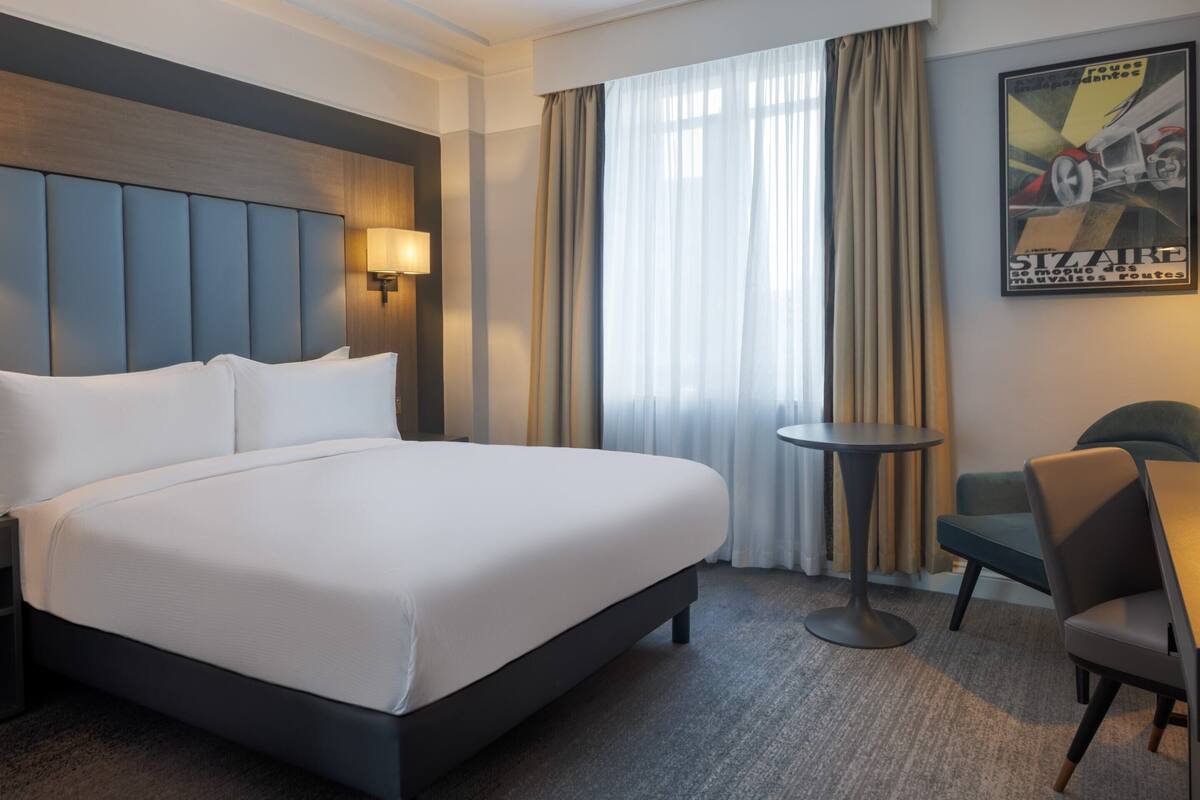 Une chambre d’hôtel avec un grand lit, une table de chevet, une chaise, une petite table ronde et une affiche accrochée au mur.