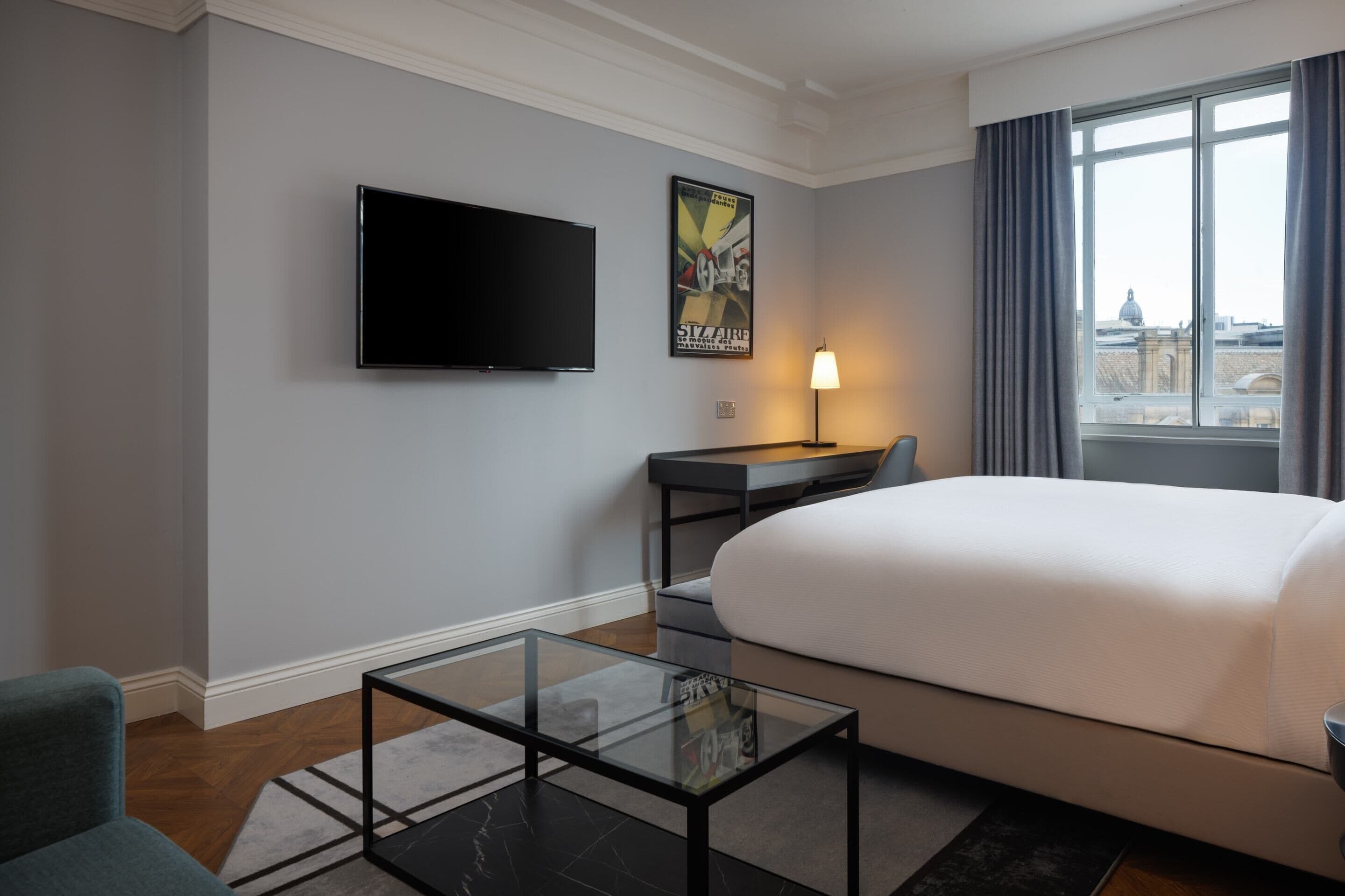A hotel room with a bed, a desk, a television, a glass coffee table, and a window with curtains.