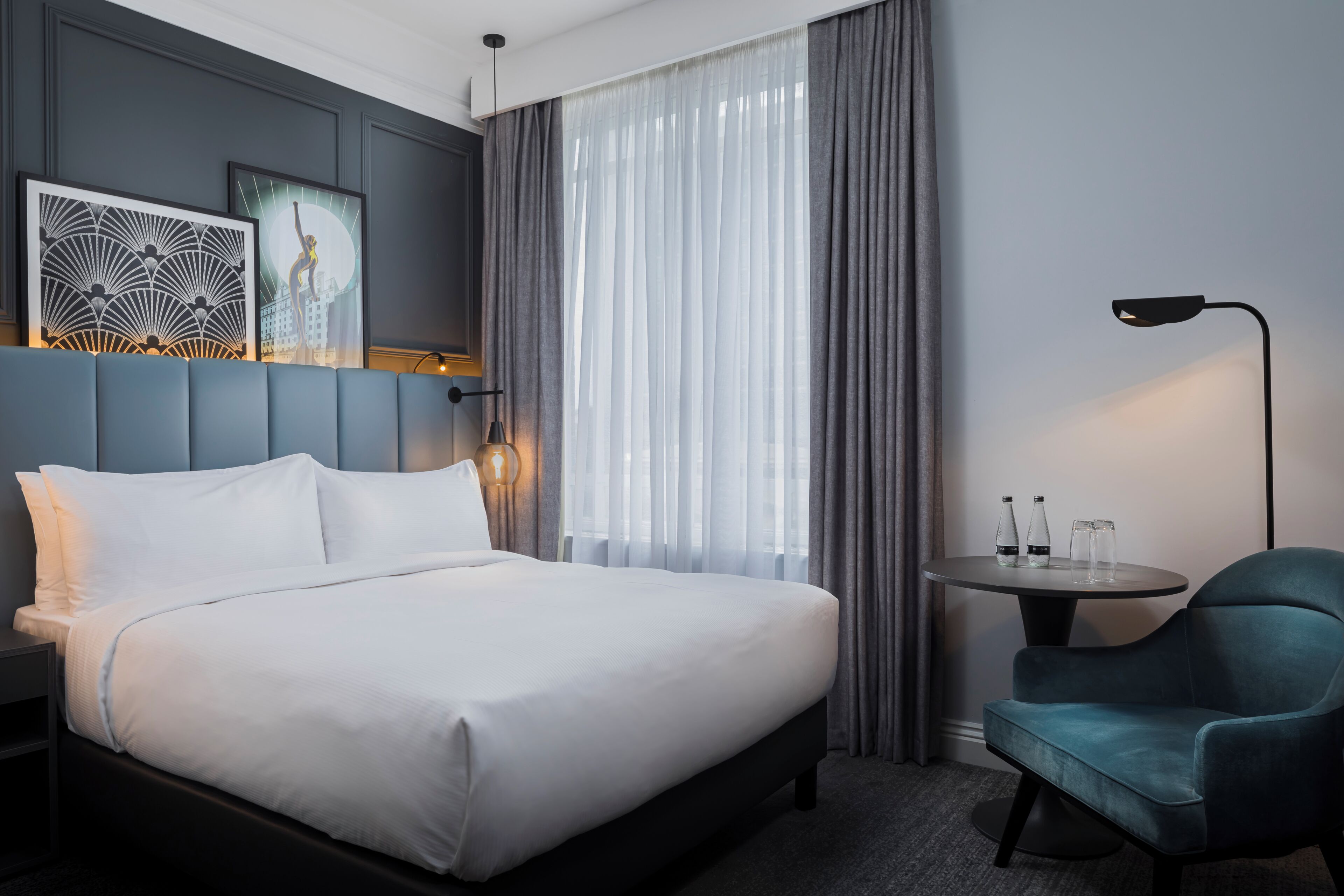 A hotel room with a bed, a chair, a table with glasses, and artwork on the wall.