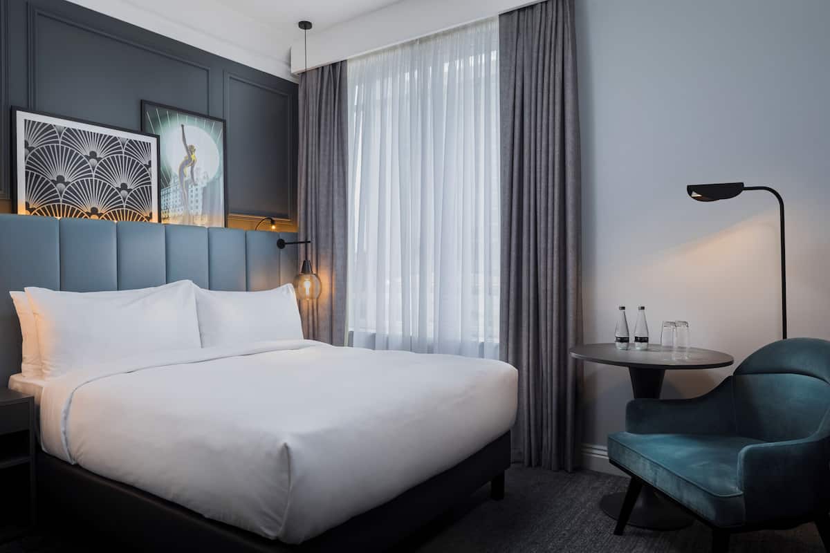 A hotel room with a bed, a chair, a table with glasses, and artwork on the wall.