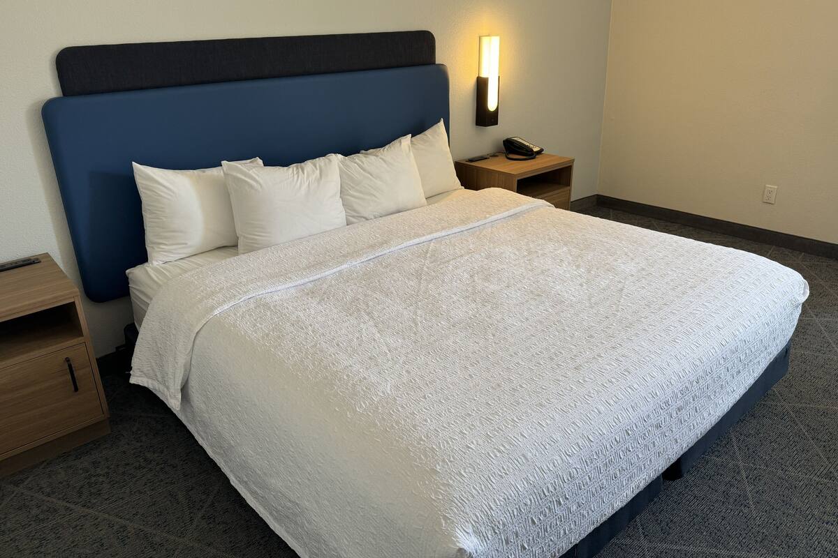 A neatly made bed with a blue headboard, white pillows, and a white textured bedspread. There is a bedside table with a lamp and a phone on it. The room has a carpeted floor and a plain wall.