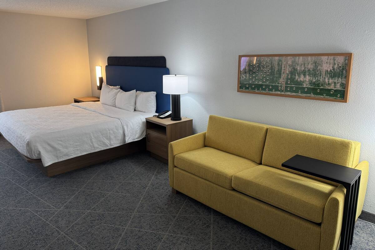 A hotel room with a bed, a sofa, a nightstand, a lamp, and a painting on the wall.