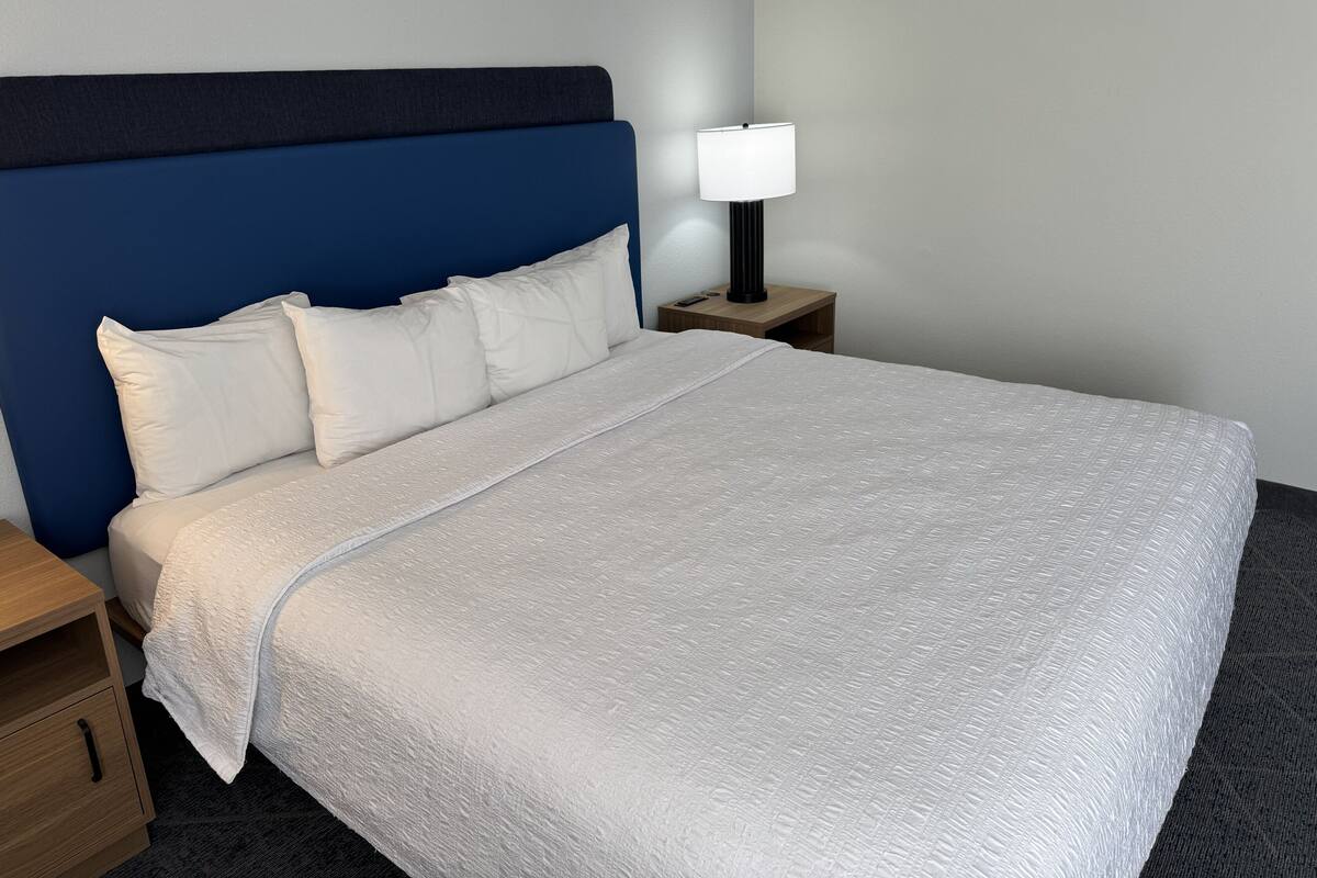 A neatly made bed with a white duvet and pillows, a blue headboard, a bedside lamp, and a wooden nightstand.