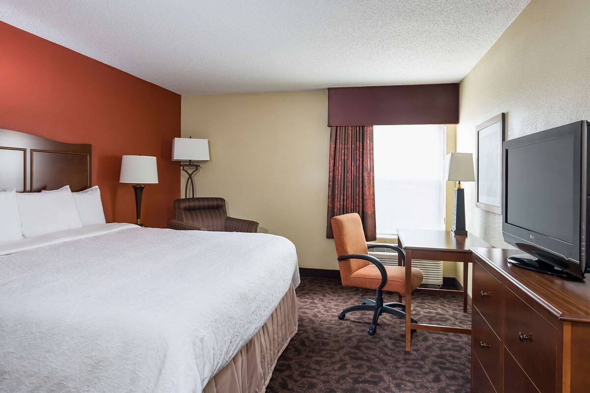 A hotel room with a bed, a desk with a television, a chair, and a lamp.