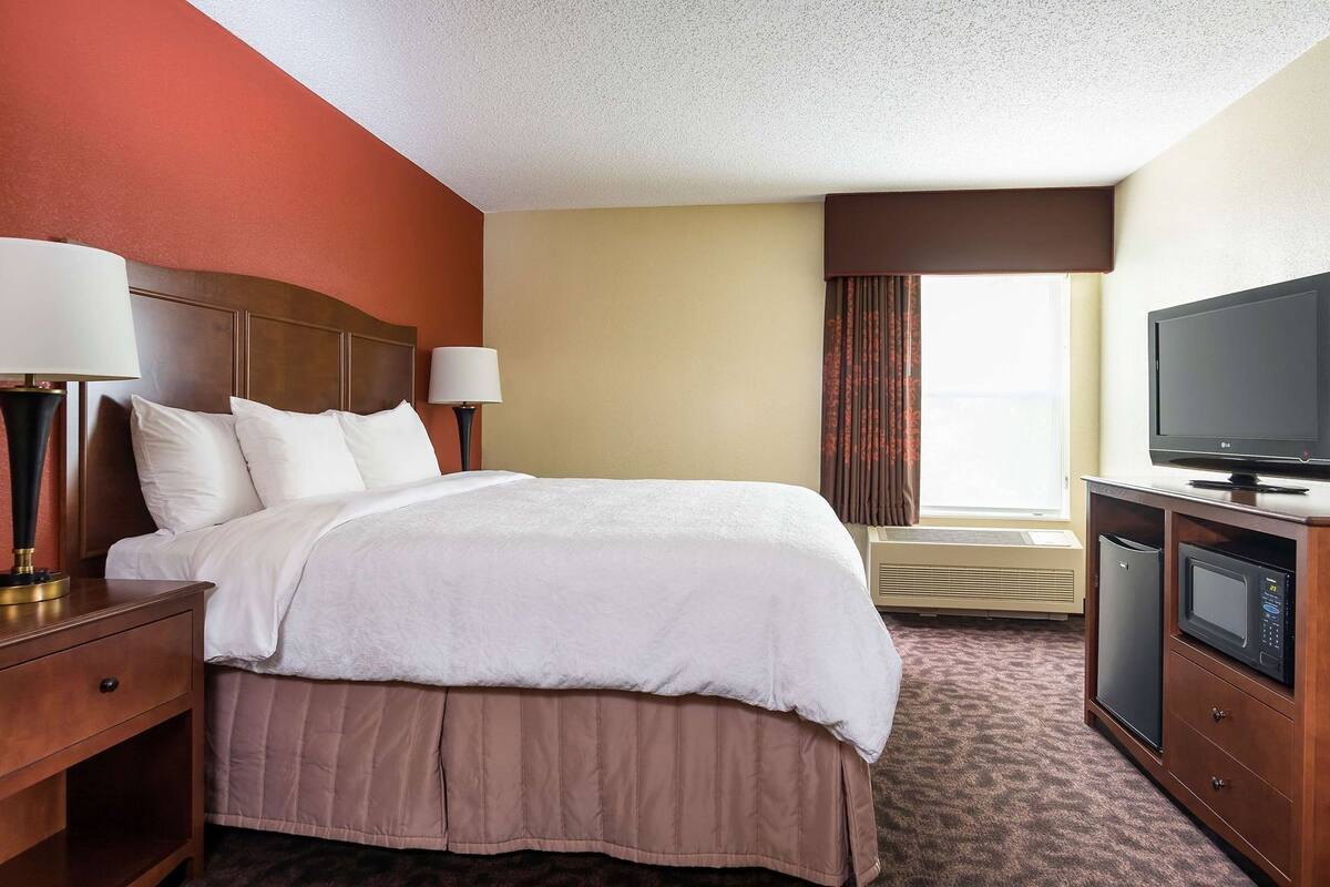 A hotel room with a bed, bedside lamps, a television, an air conditioning unit, and a window with curtains.