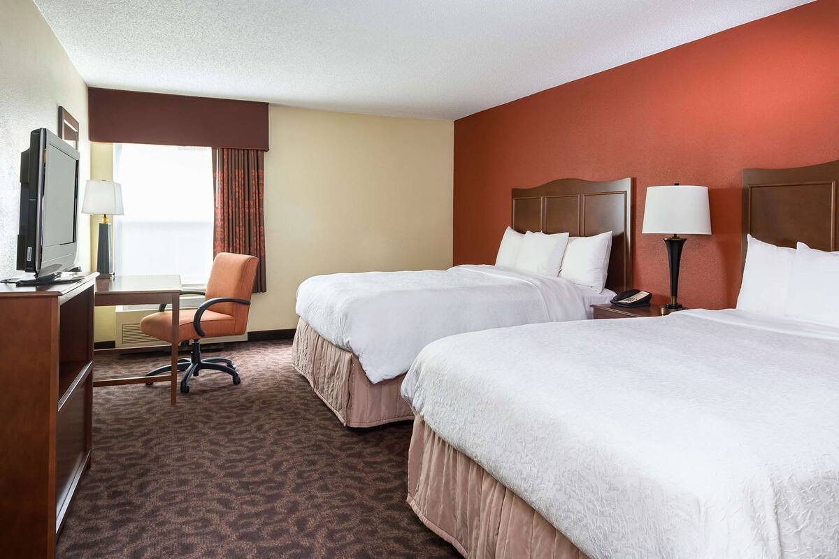 A hotel room with two beds, a desk with a TV, a chair, and a window with curtains.