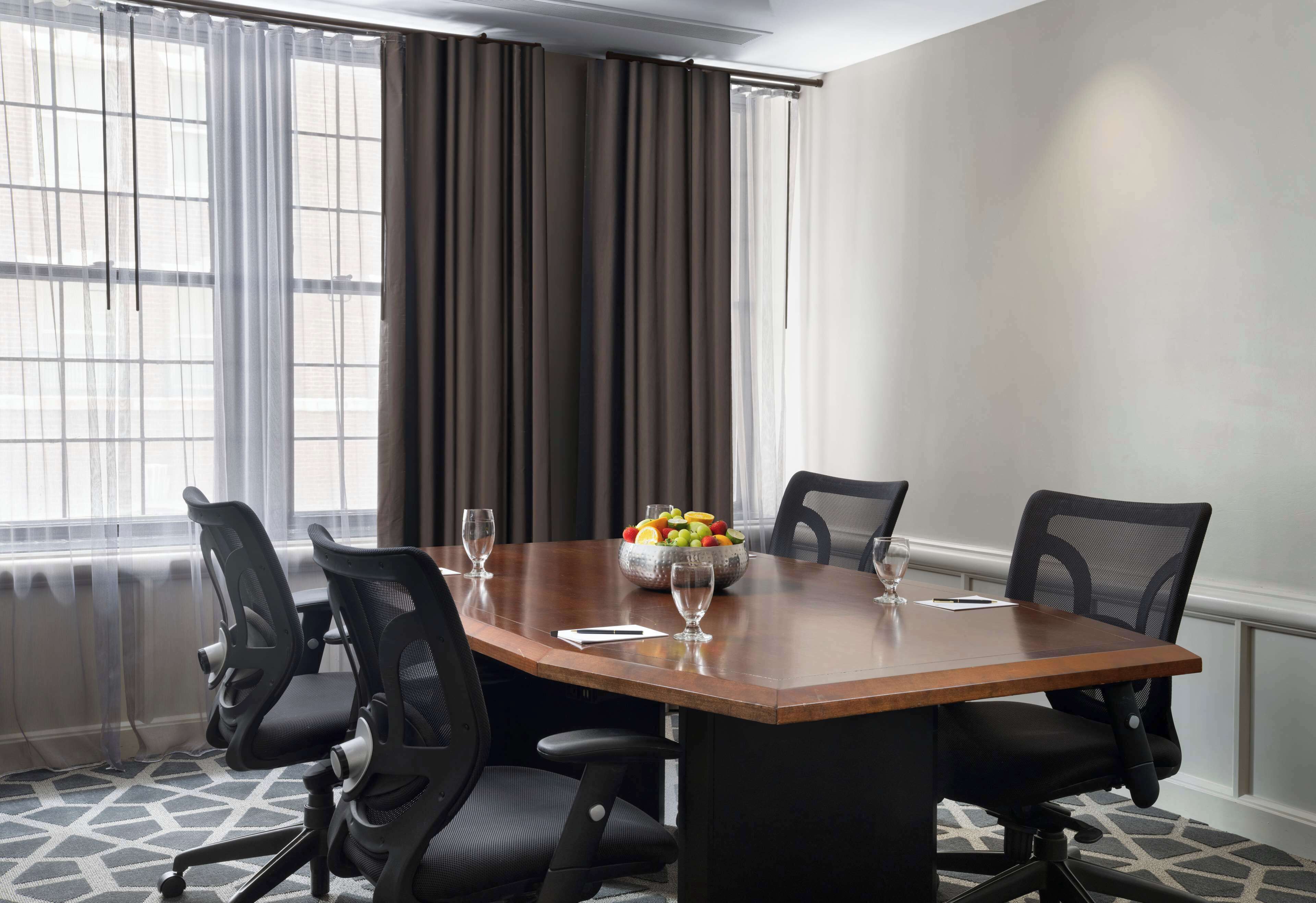 A conference room with a wooden table, four black office chairs, a fruit bowl, and two wine glasses.