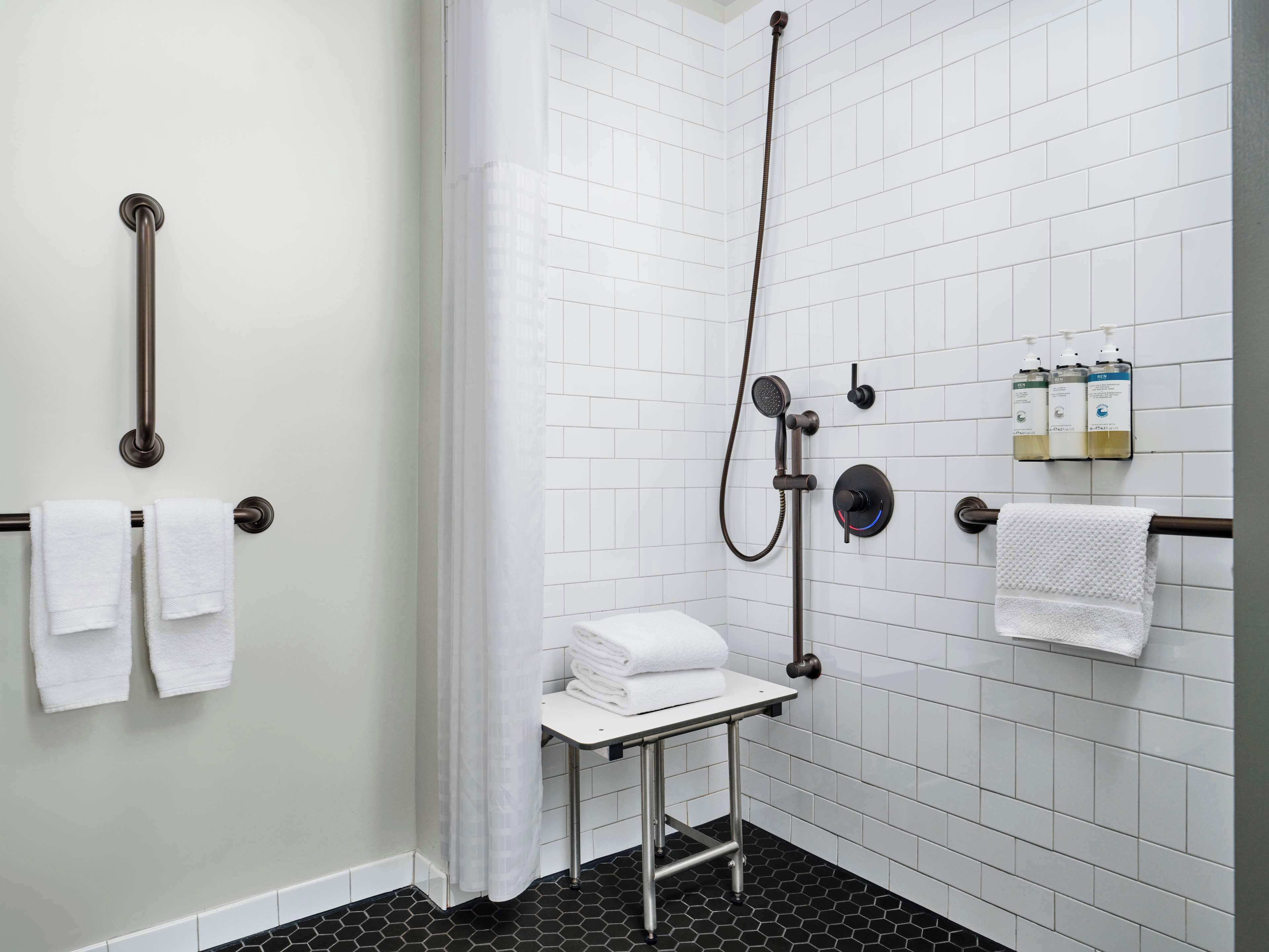 Un baño moderno con paredes de azulejos blancos, suelo hexagonal blanco y negro, ducha con cabezal de ducha manual, toallero y una pequeña mesa con toallas dobladas.