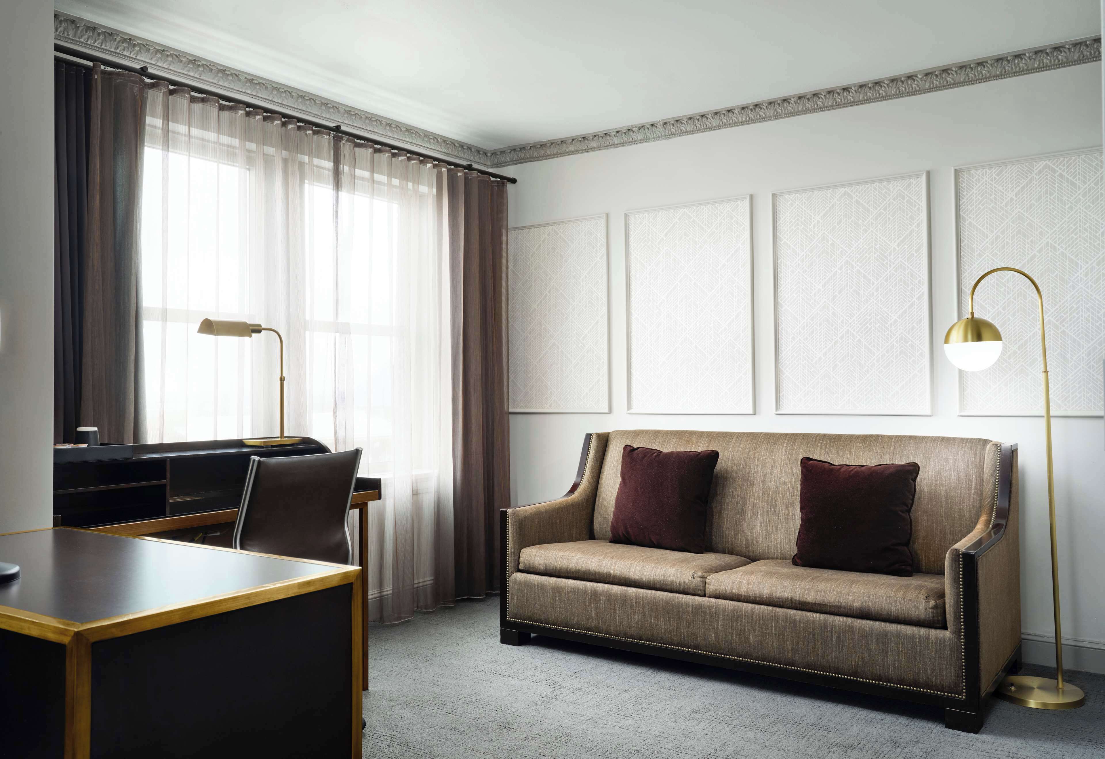 A hotel room with a sofa, a desk, a chair, and a floor lamp.