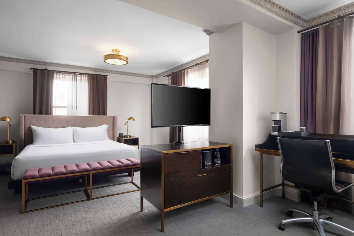 A hotel room with a bed, a TV, a desk with a chair, and a bench.