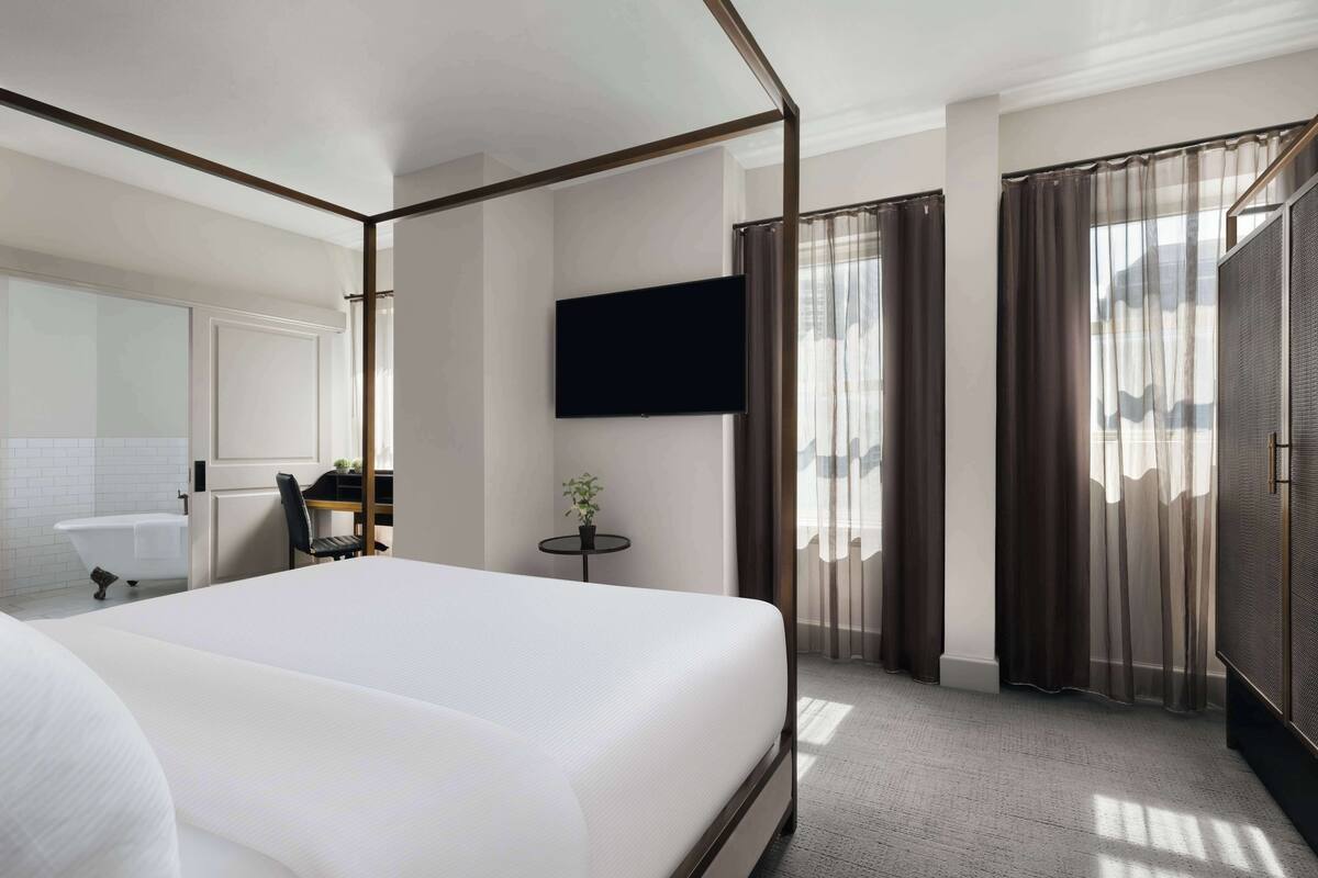 A modern hotel room with a large bed, a flat-screen TV, a small table with a plant, and a bathroom with a bathtub and toilet.