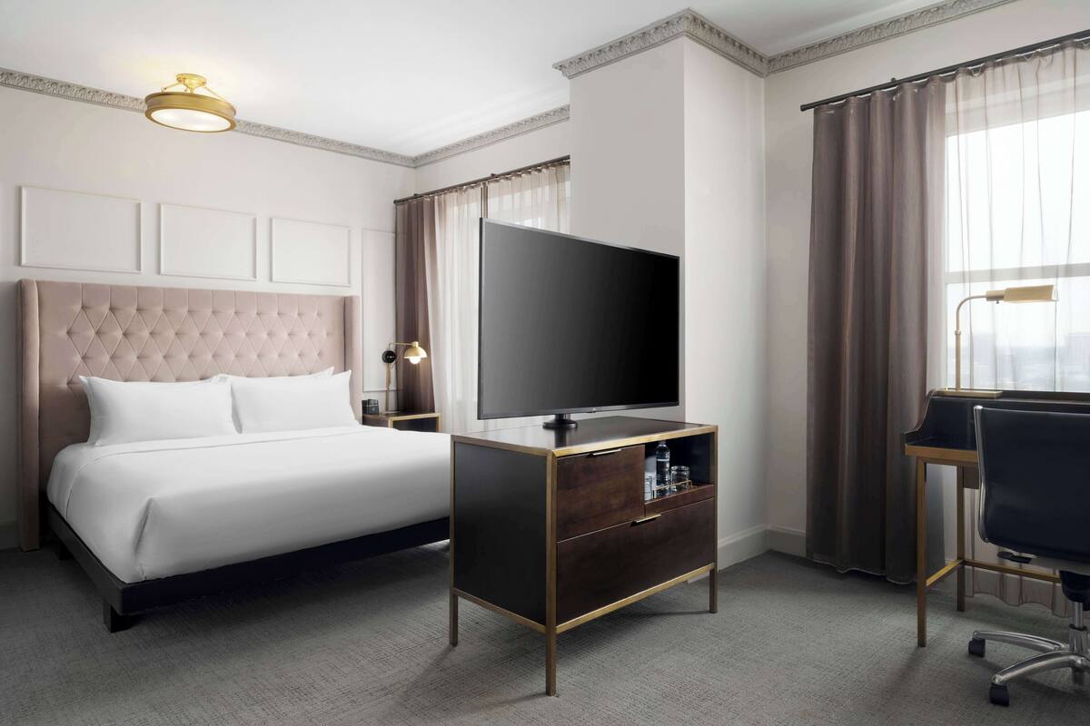 A hotel room with a bed, a TV on a wooden cabinet, a desk with a chair, and a large window with curtains.