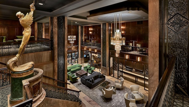 A spacious lobby with a grand staircase, a statue, a seating area with green sofas, and a well-lit reading area with bookshelves.