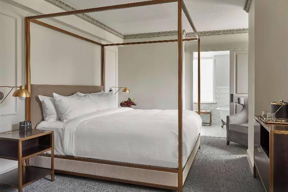 A four-poster bed with a wooden frame, a bedside table with a lamp, and a chair in a hotel room.