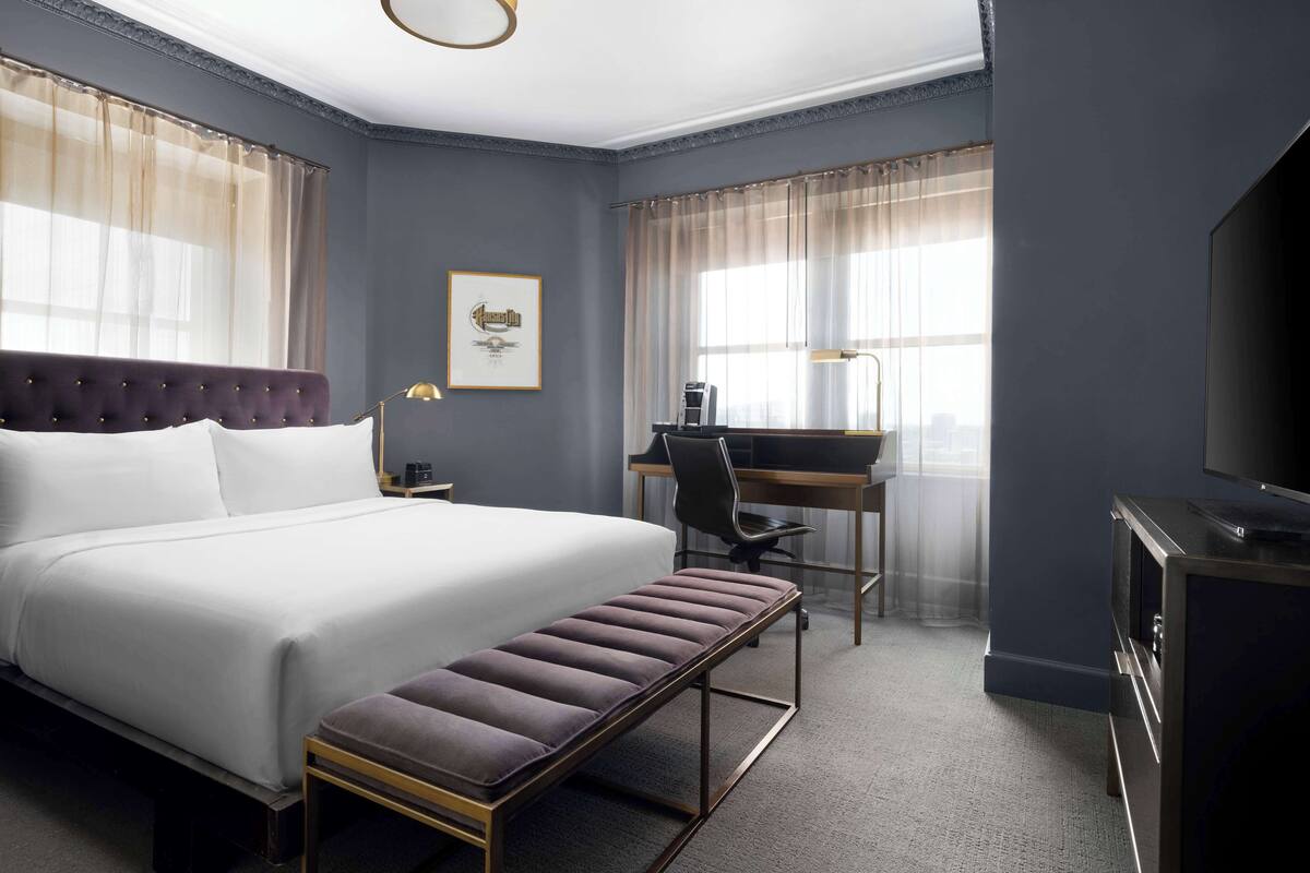 A hotel room with a large bed, a desk, a chair, a bench, and a TV.
