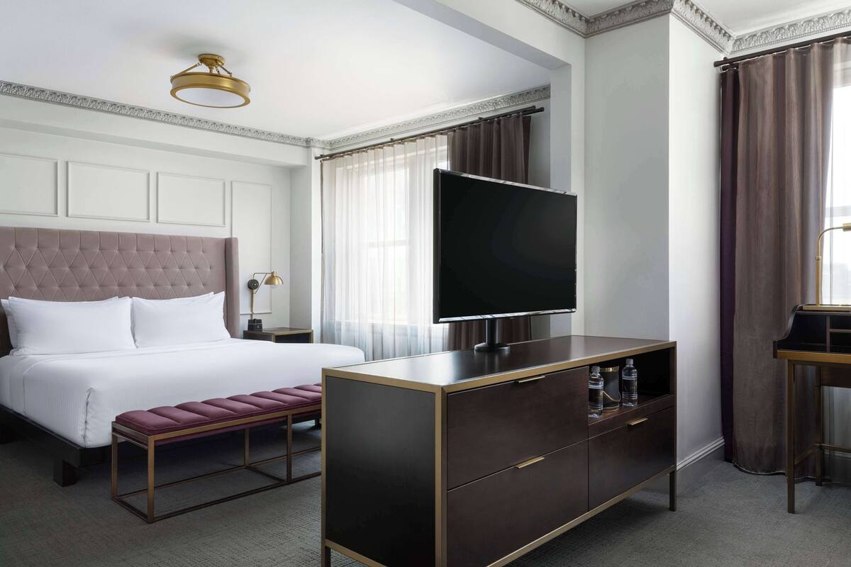 A hotel room with a large bed, a flat-screen TV, a wooden cabinet, and a bench.