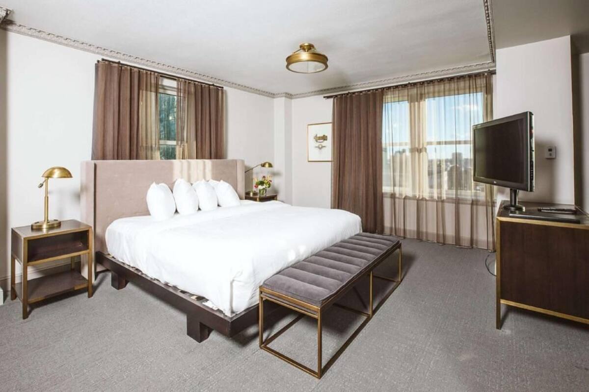 A hotel room with a large bed, a bench, a TV, a nightstand, and curtains.