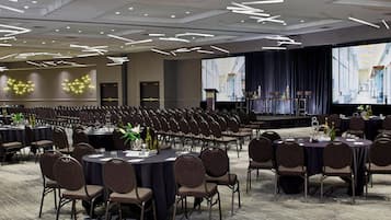 A conference room with rows of chairs, tables with centerpieces, and a stage with a large screen.